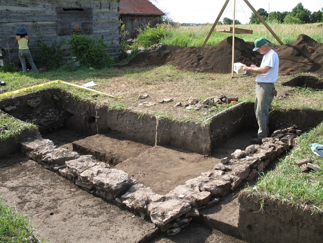 A Caucasian male archeologist stands in an archeological excavation that has revealed a fieldstone foundation and takes notes on a piece of paper.