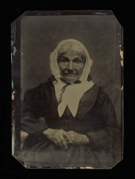 Historic black and white photo of an African American woman seated looking past the camera. She is wearing a dark-colored dress with a white bonnet around her head.