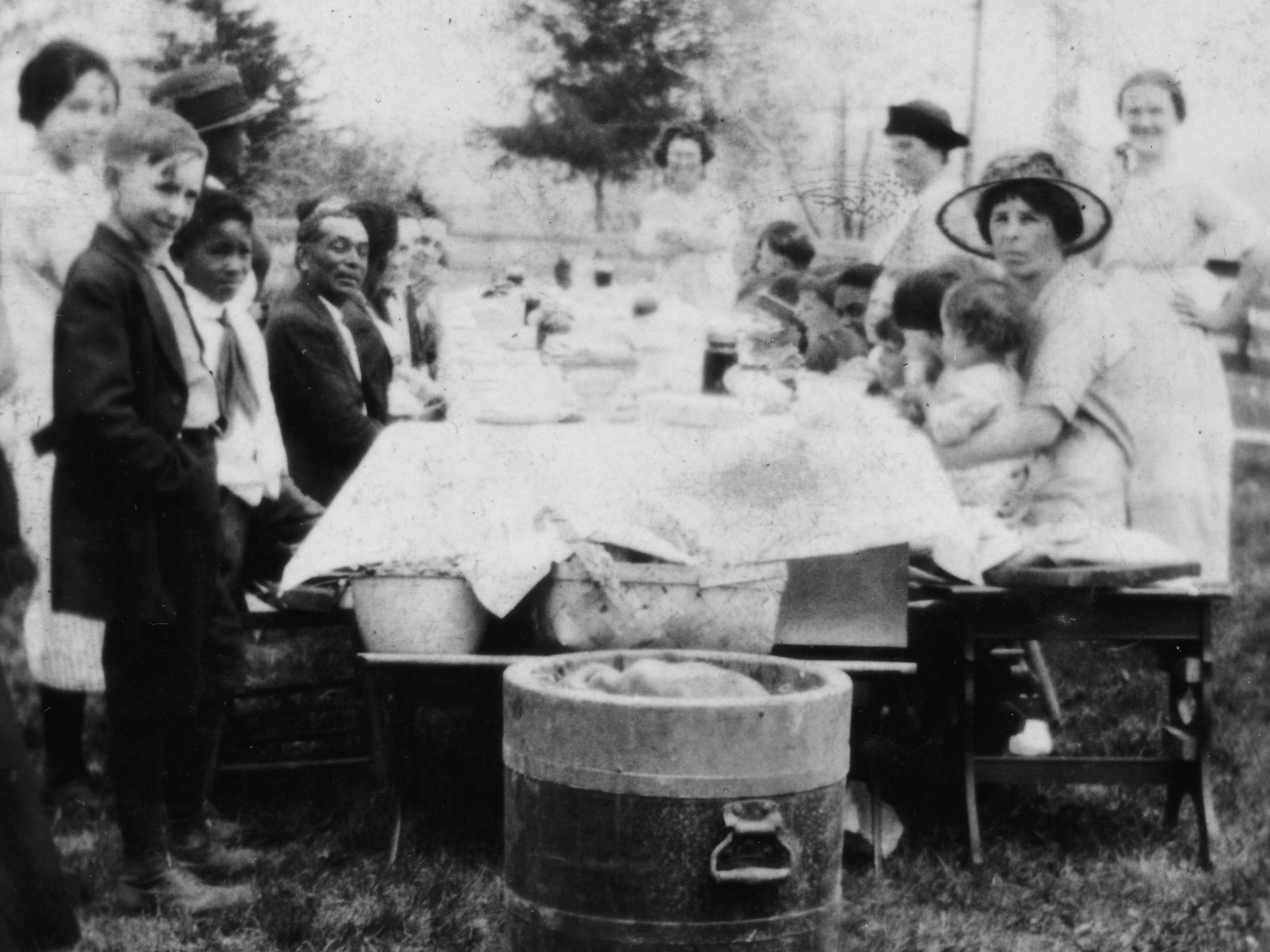 You are looking down the length of a long table with food and baskets on top. A mixed group of African American and European American people sit and stand along the sides. They are looking at you and smiling.