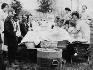 You are looking down the length of a long table with food and baskets on top. A mixed group of African American and European American people sit and stand along the sides. They are looking at you and smiling.