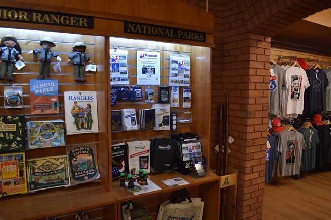 A metal stand on a gift shop counter holds seven rubber stamps. Passport stamp stickers, patches, and books hang on the wall behind it.