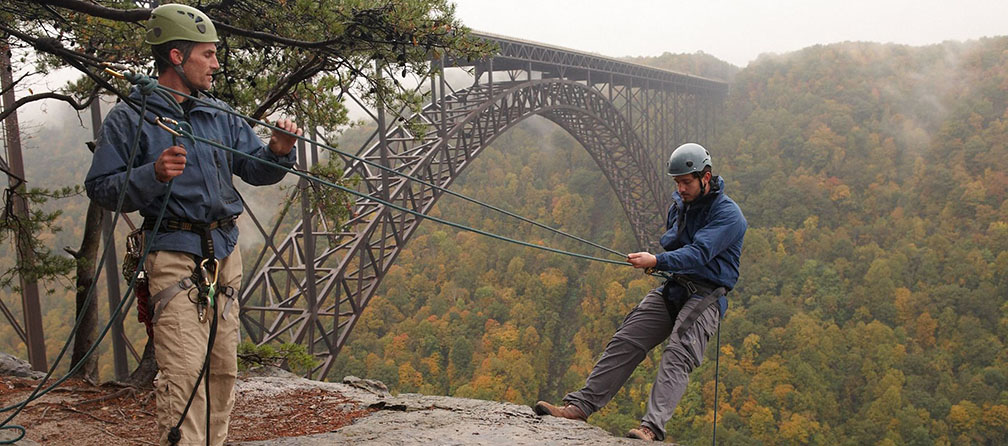 two rock climbers on a ledge overlooking a large arched bridge spanning a deep river gorge