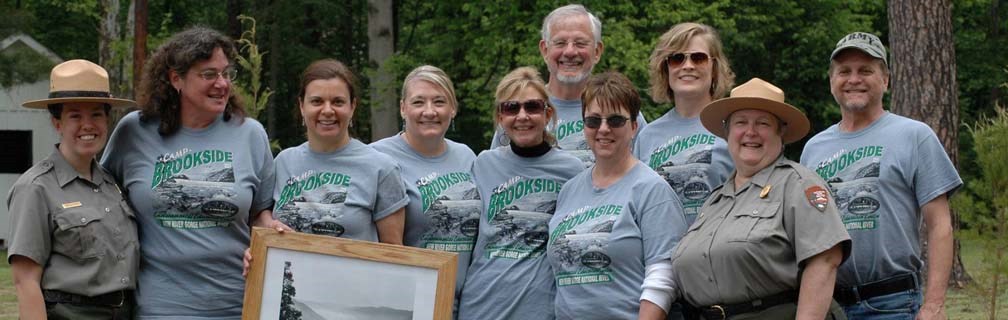 A group of people standing with two uniformed park rangers. One of the people is holding a framed historic photograph of the camp.