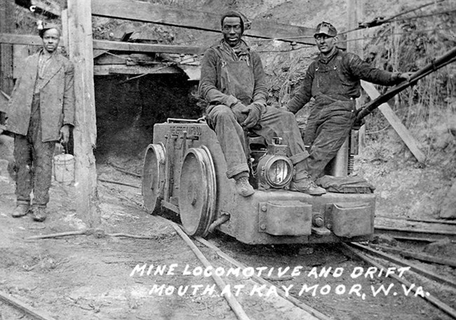 Three miners sitting on a mine cart on tracks coming out of a mine entrance
