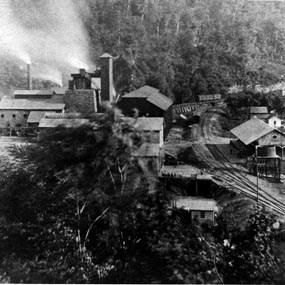 Large buildings with large chimneys spewing smoke on the side of a hill next to railroad tracks.
