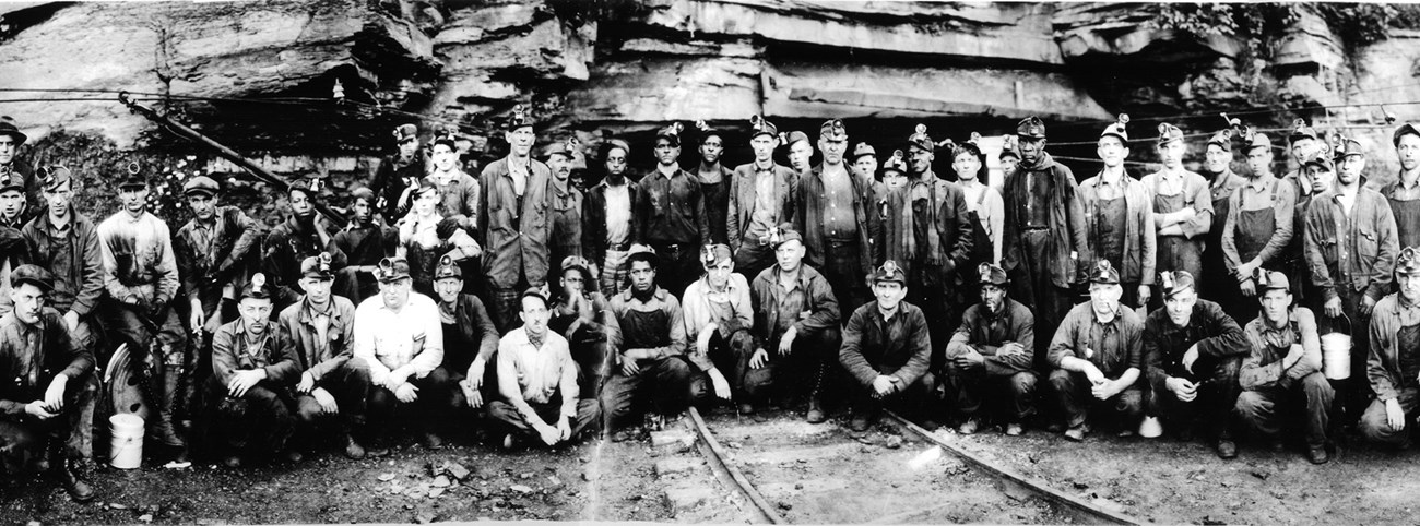 About 50 men of various ages and races wearing mining clothes and headlamps standing in front of a mine entrance