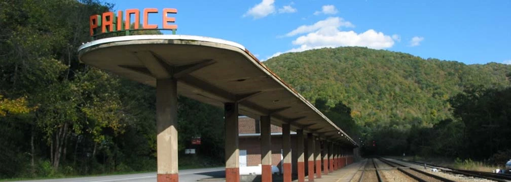 Prince Station Today Covered railroad platform with an attached station that says Prince next to railroad tracks with green mountains in the background.