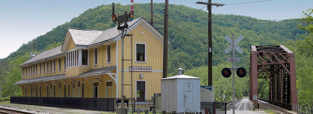A long yellow wooden building next to railroad tracks with a metal railroad bridge next to it