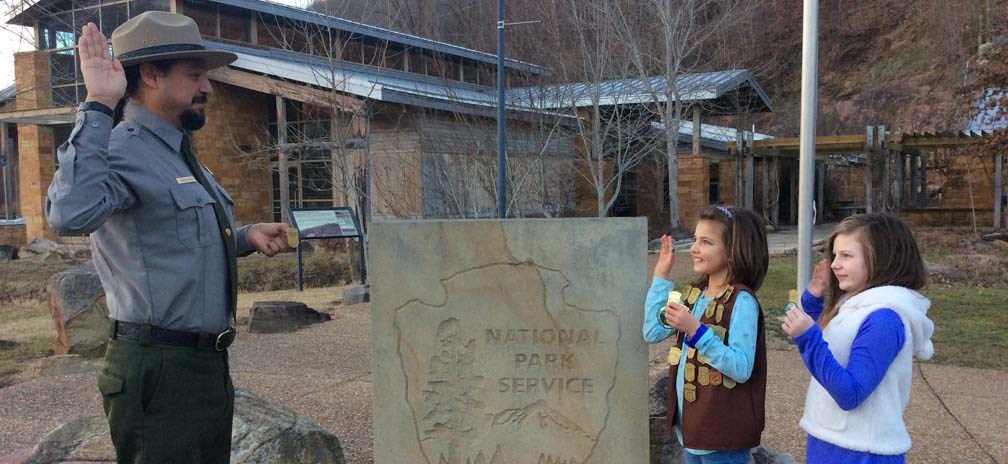 Two girls and a ranger with their right hands raised in front of a national park sign as they take the Junior Ranger pledge