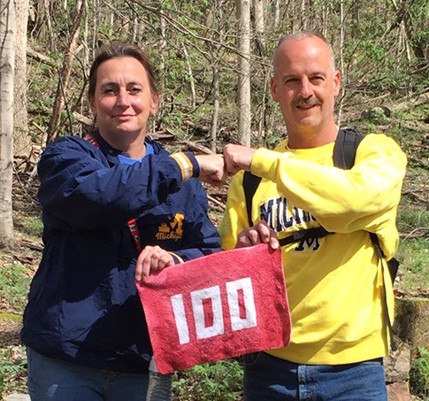 Two hikers fist bump as they hold a towel between them with "100" on it.