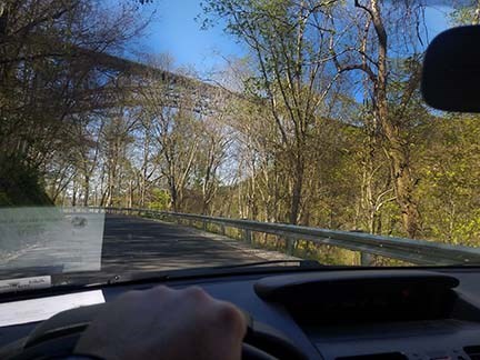 A person inside a car driving a winding road towards underneath an arched bridge