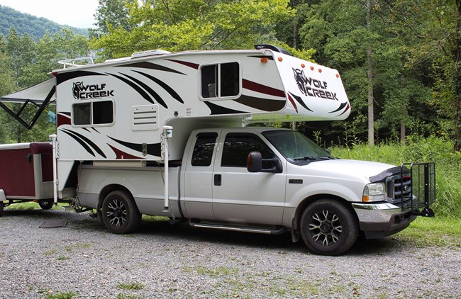 A white pickup truck with a red and white truck camper on it parked in the gravel at a grassy campsite