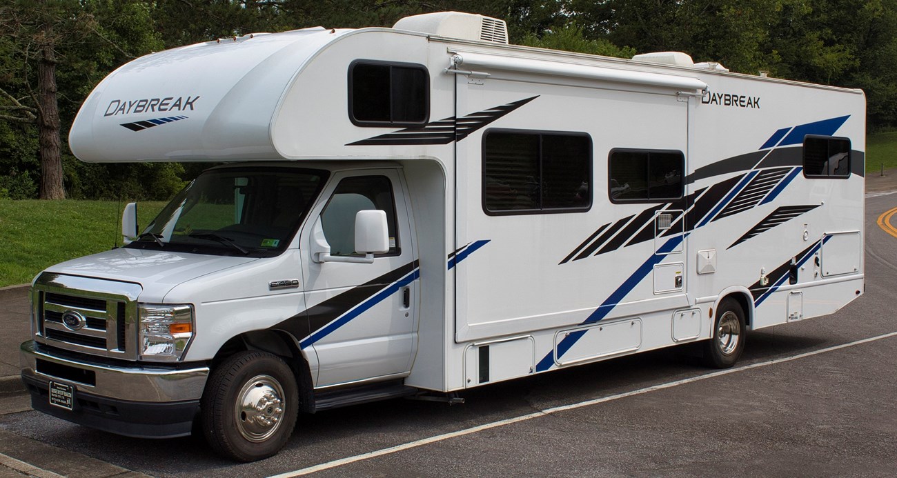 A long white and blue RV parked in a parking lot in front of green trees