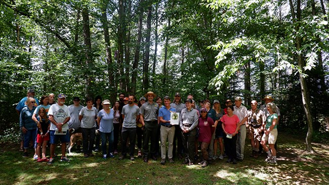 A group of about 34 people posing in front of trees in a forest. In the middle of the group are 3 park rangers and a person holding a white and green certificate