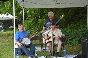 A man with a banjo, a man with a guitar and a woman with a bass are playing music under a canopy.