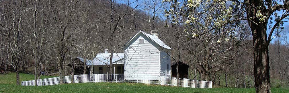 Color photo of a green hill with a white wooden farmhouse in the center. Small wood outbuildings flank the house and a white picket fence surrounds them all. Behind all is a forested hill with a blue sky.