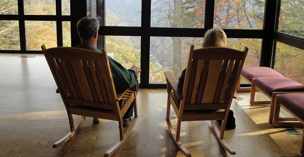Two visitors sit in wooden rocking chairs, looking out the back window of the Visitor Center at the New River.