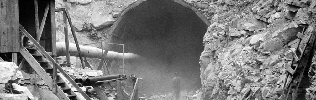 A circular tunnel cut into rock with a pipe coming out of it and a worker walking into it