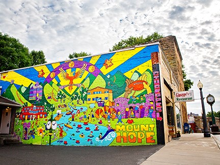A colorful mural on the side of a building with images of the river, buildings, people, mountains and rays of sunshine.