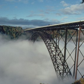 A reddish brown metal arch bridge across a mist filled gorge
