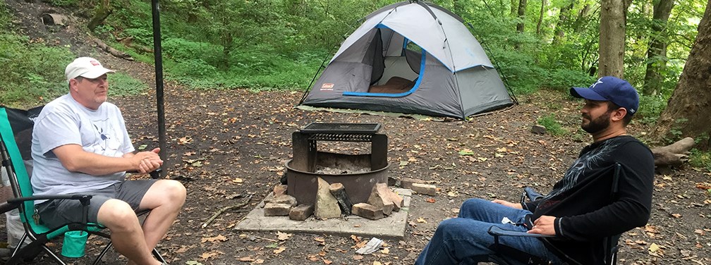 Two male campers sit in camping chairs on either side of a fire pit with a tent in the background