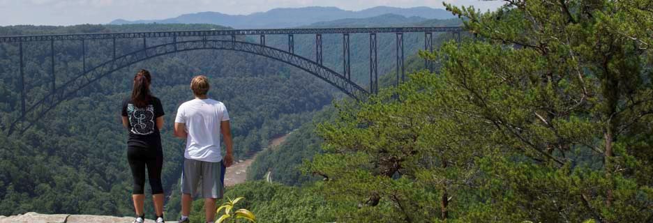 Colored photograph of two hikers, overlooking the gorge, facing towards the New River Gorge Bridge in the distance.