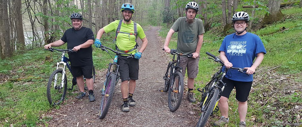 A colored photograph of four bikers, wearing helmets, standing on the trail, next to their bikes.