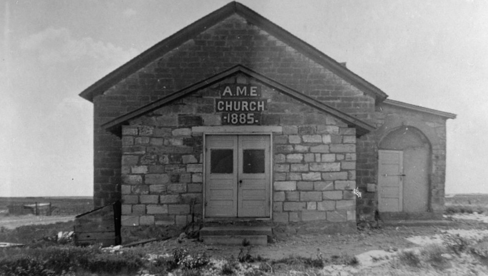 Black and white photo church on the prairie, made of large limestone bricks. At the front of the church are white double-doors. Above these doors are the words "A.M.E. CHURCH 1885".
