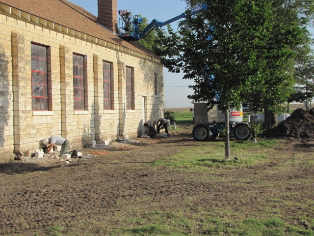 Crew members work on bricks at the base of a limestone building with large framed windows. A tree is in front of a lift machine that supports a man inspecting the building's chimney. The ground has tire tracks all over it.