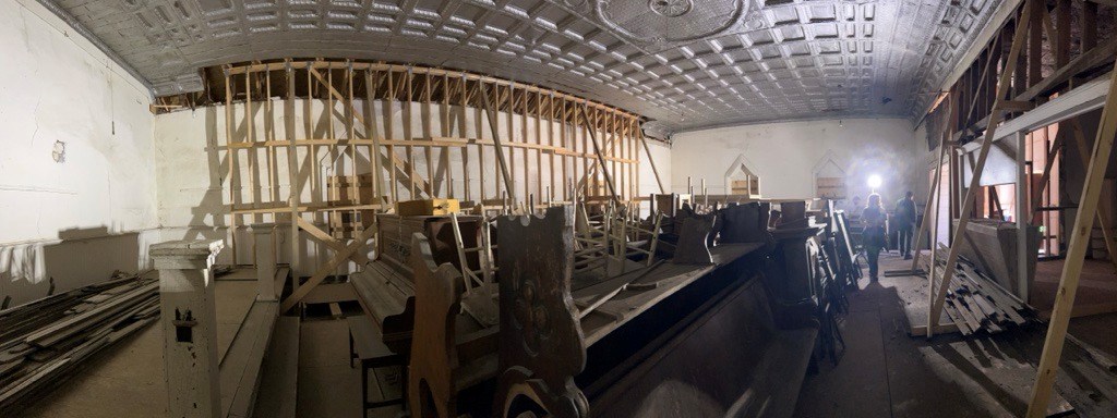 Panoramic photo inside old building. A bright lamp on the right lights the inside. Several stacks of pews and chairs are in the middle. Lumber is stacked on a raised platform to the left. Two long walls are held up with wooden supports.