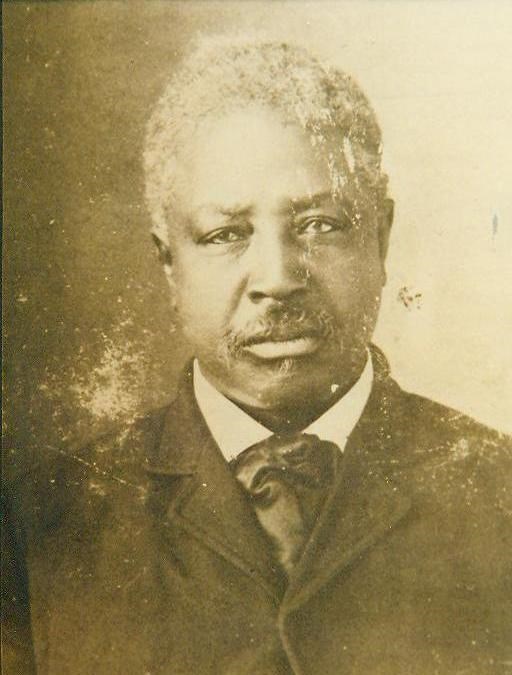 A sepia photo of a man from the chest up with short white hair and wearing a dark suit.