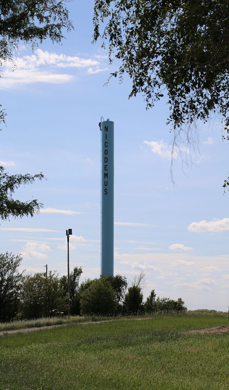 The vertical column of the Nicodemus Water Tower stands across a grass lawn.