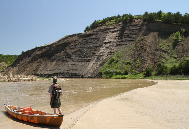 A canoer stands on shore of a river by his boat and looks at an exposed bluff of dark sand and clay soil