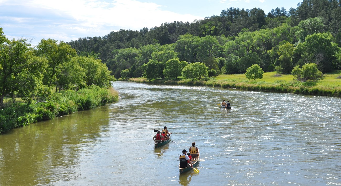 People in three canoes float down a broad stretch of the Niobrara river, with trees to the sides