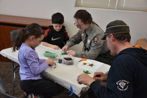A park ranger teaches people how to make a turtle out of craft beads at Kids' Craft Day. A park ranger shows two children and an adult how to make a craft turtle out of beads.