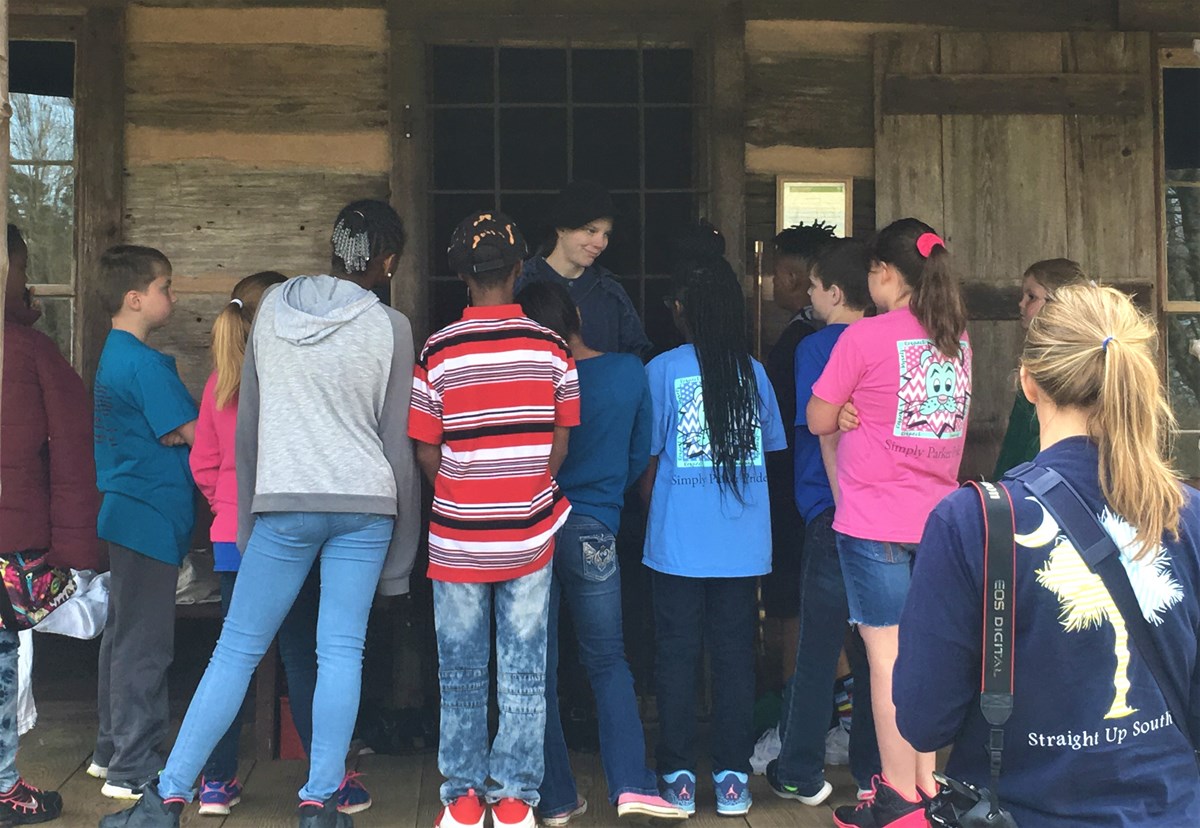 A group of children gather around a living historian on the porch of the cabin