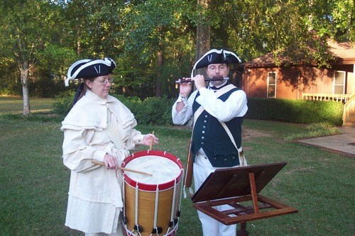 Two people in 18th century clothing are playing a violin and a drum.