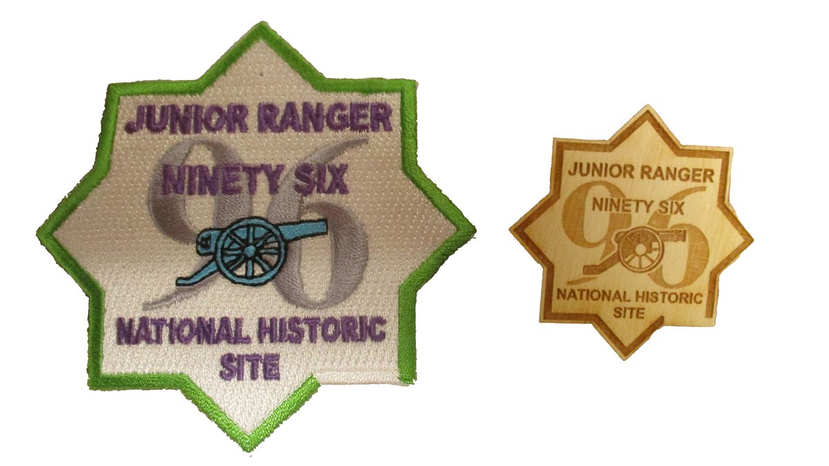 Eight-star shaped colored patch on left and wooden badge on right. The patch is white with a green edge and a gray "96" in the center. In front is a blue cannon. Above the cannon in purple "Junior Ranger Ninety Six" below "National Historic Site"