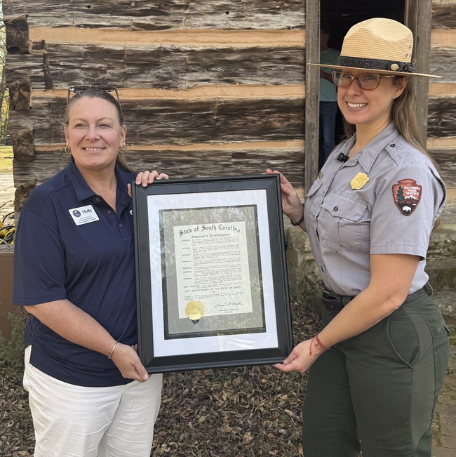 A woman in a blue shirt and white pants stands next to a park ranger. They are holding a framed document and are standing in front of a log cabin.