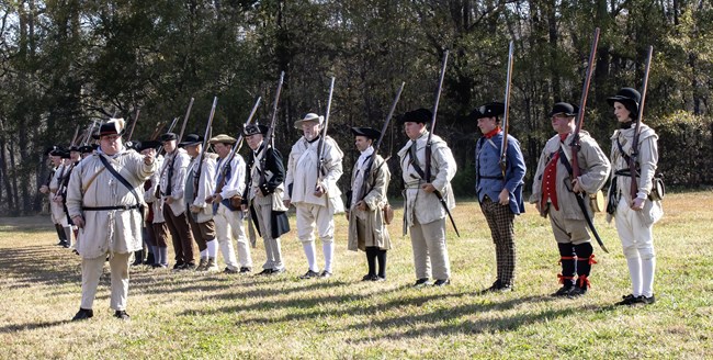Many people dressed in 18th century uniforms and carrying muskets are lined up with one man giving directions.
