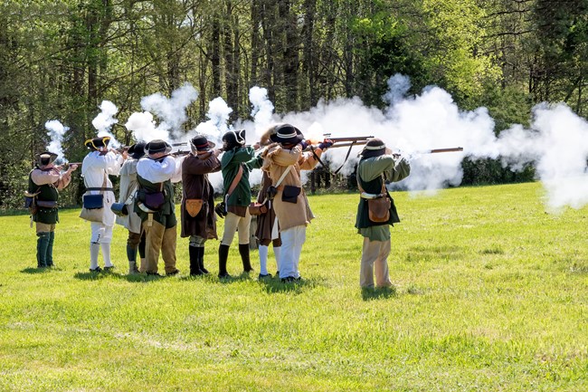 Men in 18th century clothing are firing muskets which produce a lot of gunsmoke.