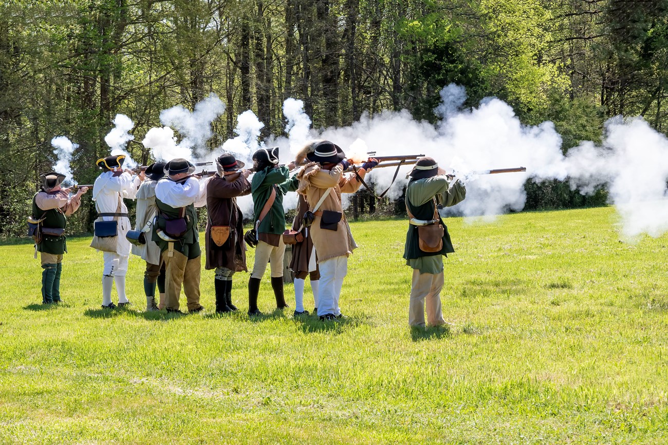 Men in 18th century clothing are firing muskets which produce a lot of gunsmoke.