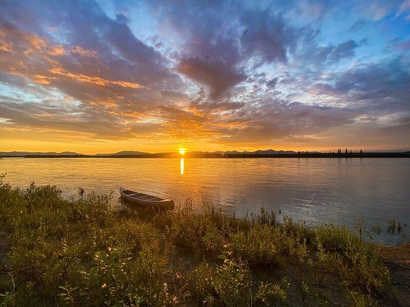 a wide body of water extends to the horizon and reflects the vibrant oranges and blues of a setting sun. A canoe in the foreground sits empty aside a bank of upright plants and grasses.