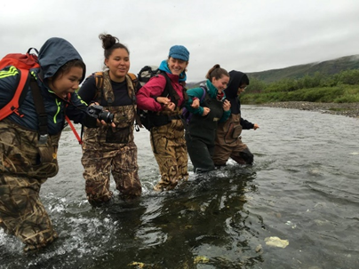 five young women link arms while crossing a body of water, each dressed in overall-like waders and warm clothing.