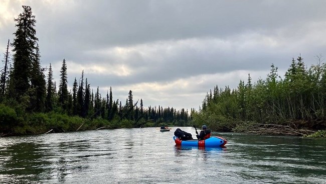 a person in a blue and red inflatable raft floats down a wide channel of tree-lined water.