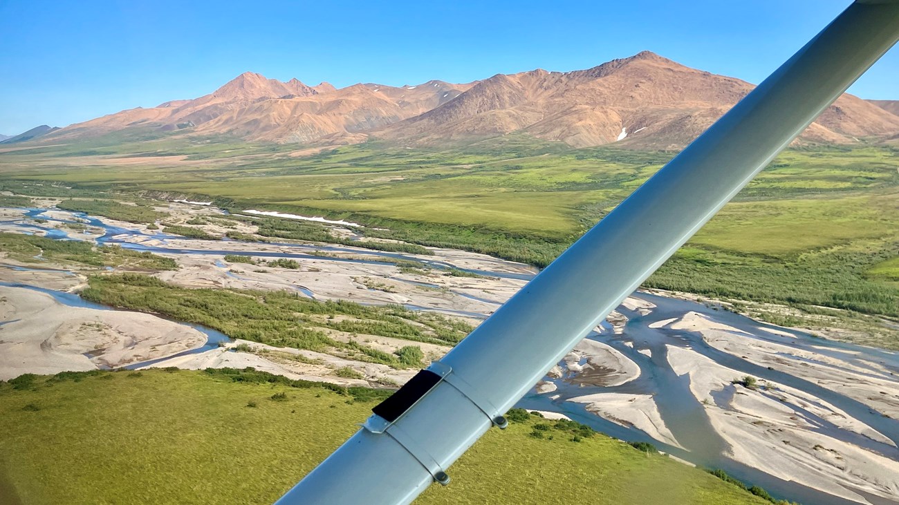 Red tinted mountains on the horizon and the braided Kelly River below the strut of Cessna 206.