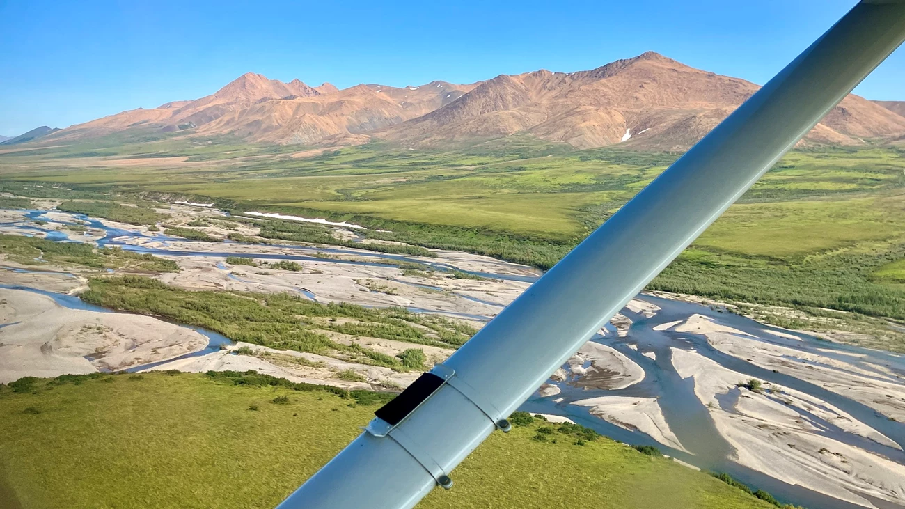Flying over the Upper Kelly River Red tinted mountains on the horizon and the braided Kelly River below the strut of Cessna 206.