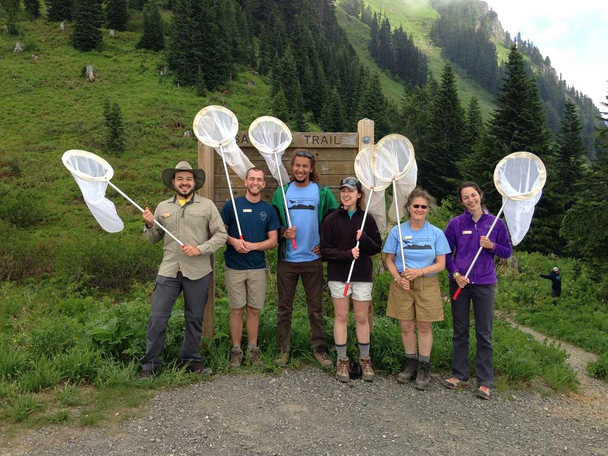 Six scientists stand in front of a sign holding butterfly nets with green trees in the background. Six scientists stand in front of a sign holding butterfly nets with green trees in the background.
