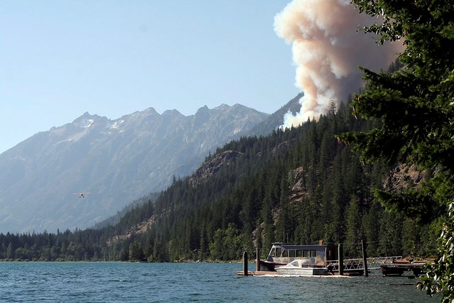 Smoke plume rises above a forested area along a lake