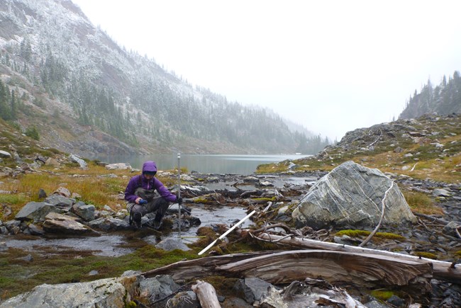 Person crouched down near small pool of water with mountains in the background